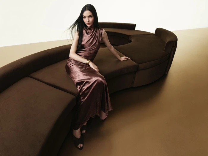 Mariacarla Boscono seated on a curved brown sofa wearing a brown satin dress for Donna Karan Fall 2025 Campaign