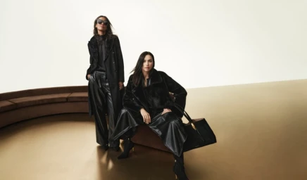 Two models in black coats posing against a curved brown bench and pale wall for Donna Karan Fall 2025 campaign.