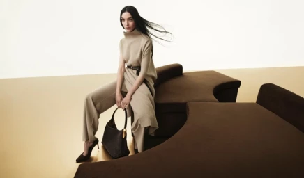 Model wearing beige knit tunic and trousers seated on a brown modular sofa holding a dark handbag.