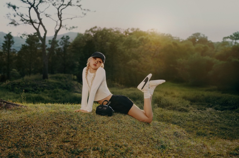 ROSÉ lying on a grassy hill wearing a white long-sleeve top, dark shorts, and white Puma sneakers in a natural outdoor landscape