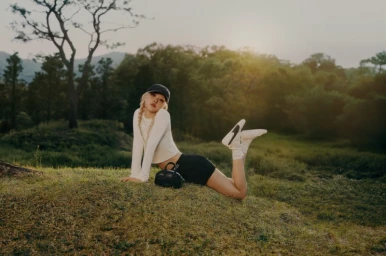 ROSÉ lying on a grassy hill wearing a white long-sleeve top, dark shorts, and white Puma sneakers in a natural outdoor landscape
