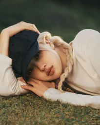 Close-up portrait of Rosé wearing a black baseball cap and white long-sleeve top while lying on grass.