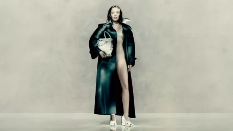 Miley Cyrus in a dark glossy coat holding a white handbag against a pale mottled backdrop for Maison Margiela