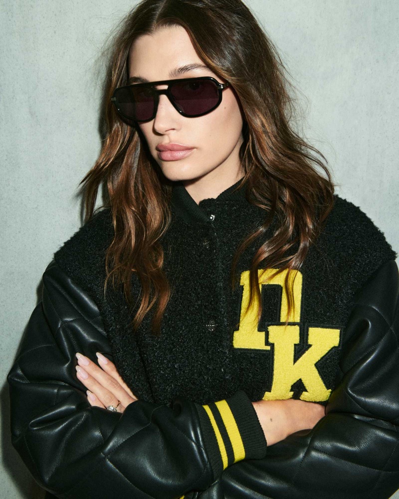 Hailey Bieber wearing black sunglasses and a black DKNY varsity jacket with yellow DK lettering against a concrete wall.