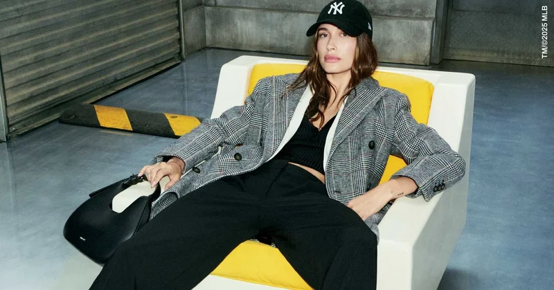 Hailey Bieber wearing a plaid blazer and baseball cap seated on a white and yellow chair for DKNY Fall 2025 campaign.