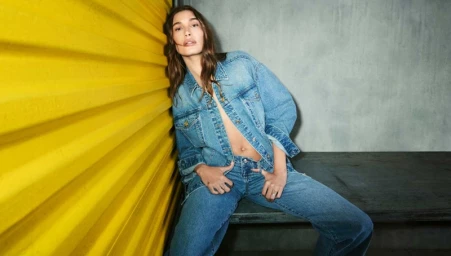 Hailey Bieber wearing a denim jacket and jeans leans against a ribbed yellow wall for the DKNY Fall 2025 campaign.