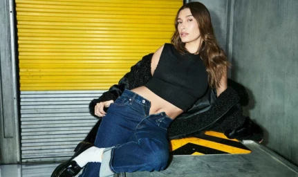 Hailey Bieber wearing a black sleeveless top, denim jeans, and a dark coat against a yellow and grey industrial background