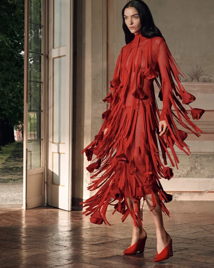 Model wearing a vibrant red fringed dress and red mules standing in a sunlit interior room near open doors.