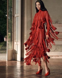Model wearing a vibrant red fringed dress and red mules standing in a sunlit interior room near open doors.