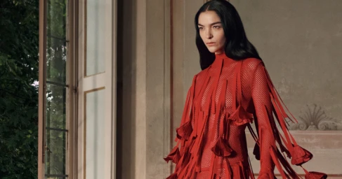 Mariacarla Boscono in a red fringed mesh dress standing indoors near a tall window for Salvatore Ferragamo