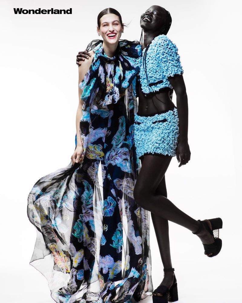 Two smiling fashion models in studio setting. The model on the left is wearing a floral patterned jumpsuit, and the right model is wearing a sky blue outfit.