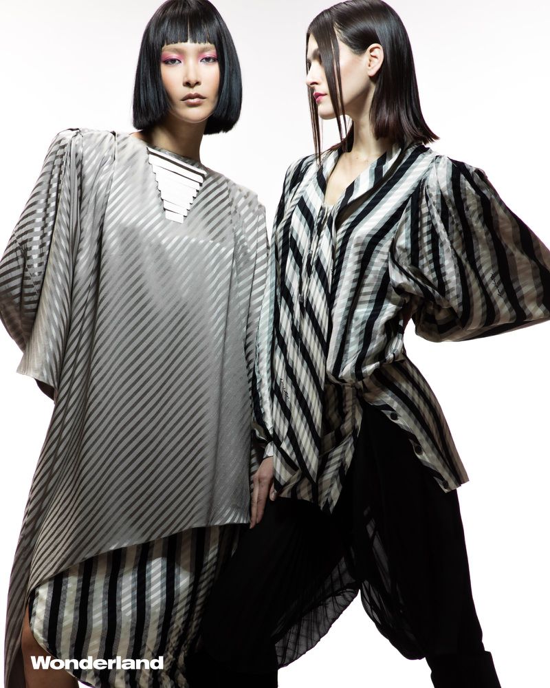 Two fashion models pose in a photo studio. One woman wears a grey and white striped dress; the other wears a black and white striped top and black pants.