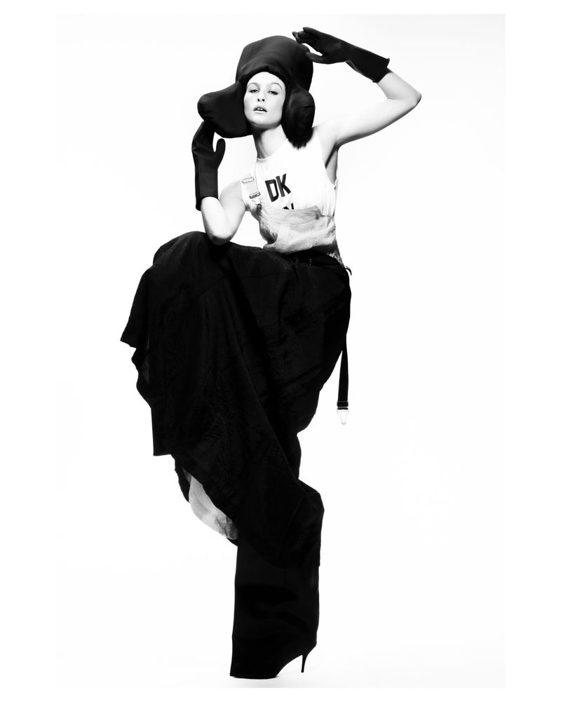 Black and white fashion photo of a woman wearing a large hat, gloves, top, and wide pants, balancing on one leg against a white backdrop.