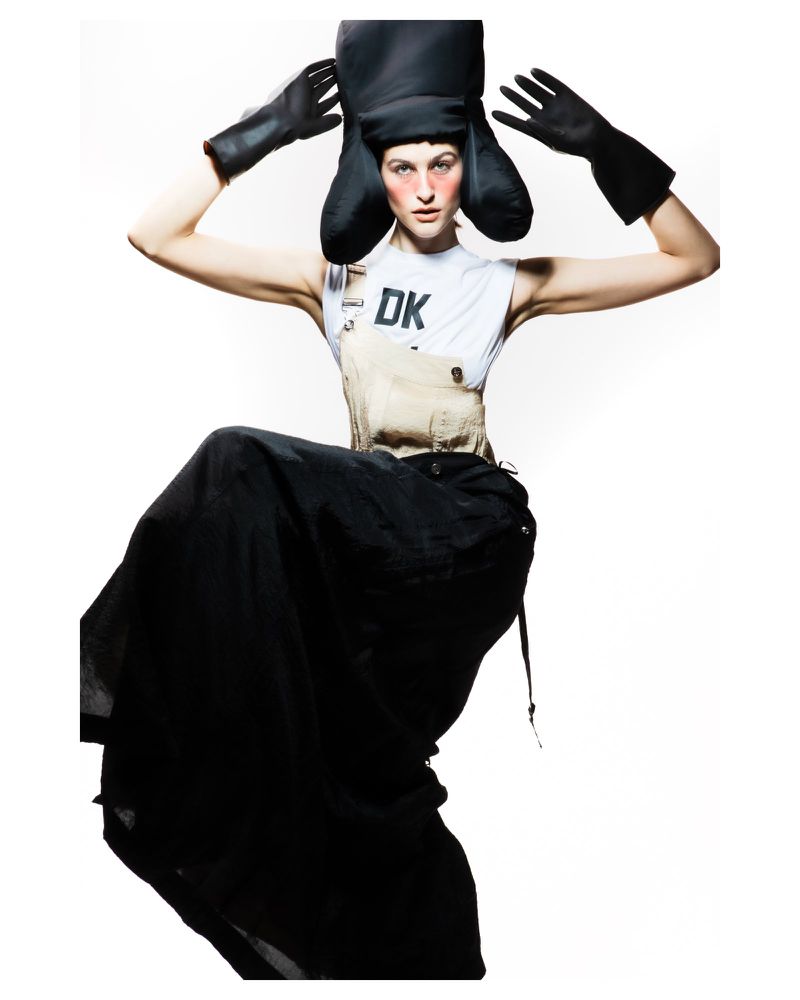 Fashion shot of a model in a white top, tan overalls, and a black skirt, wearing oversized black gloves and an unusual black headpiece.
