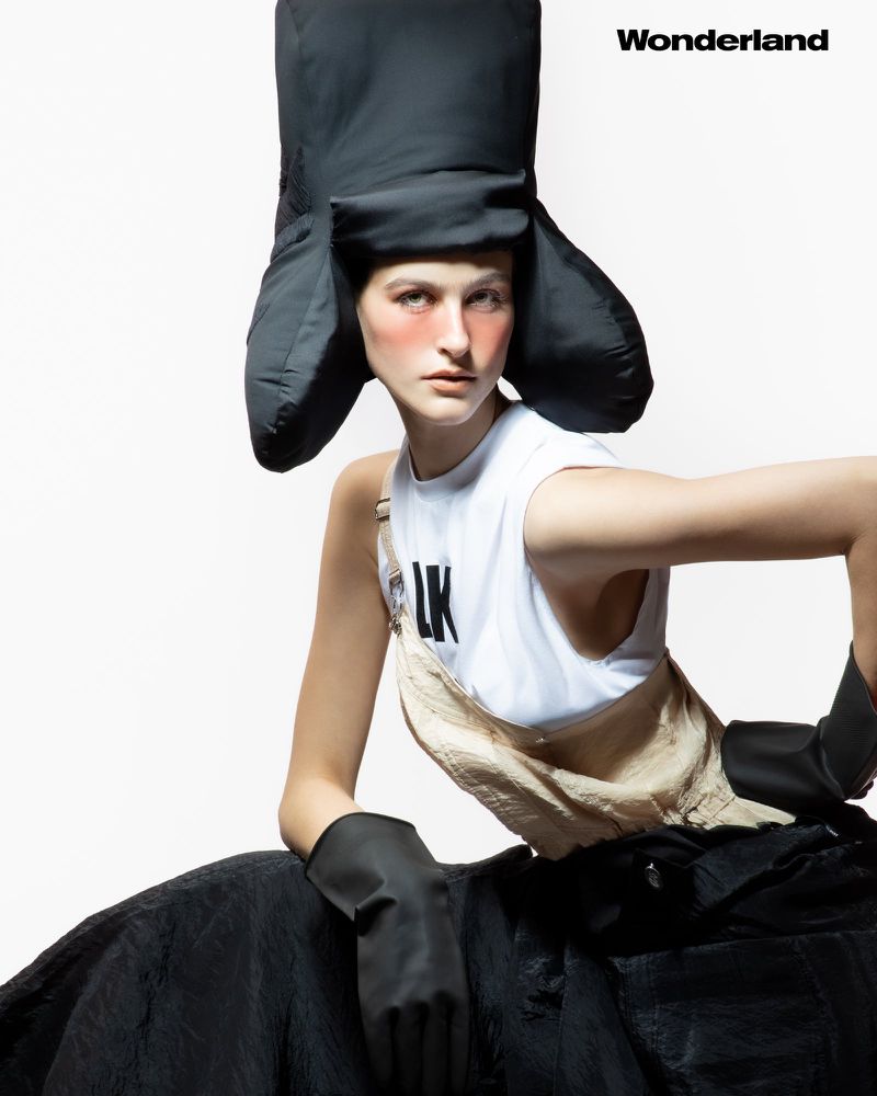 Fashion portrait of a young adult female wearing a black oversized trapper hat, white sleeveless shirt and overall dress in a photo studio.