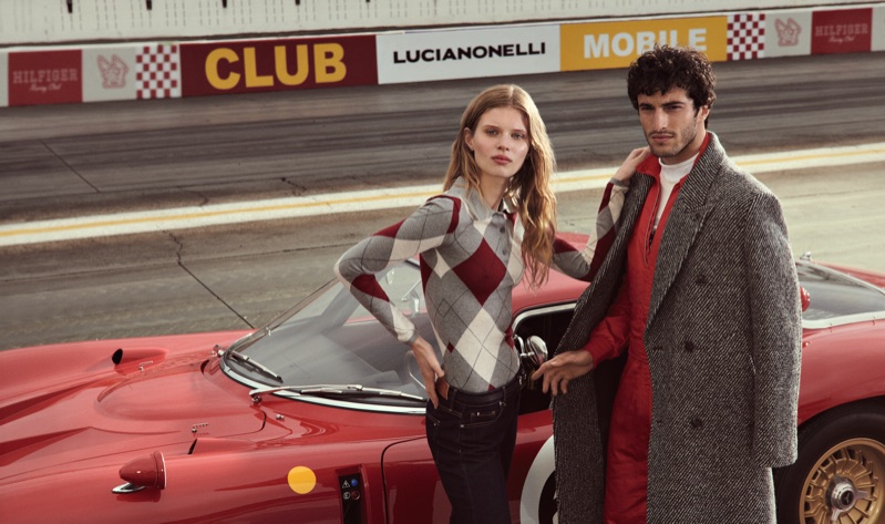 Woman in an argyle sweater and man in a red jumpsuit and grey overcoat posing beside a classic red race car on a racetrack.