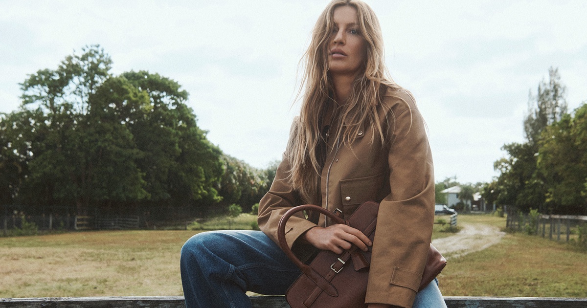 Gisele Bündchen seated on a wooden fence holding a brown leather bag, wearing a tan utility jacket and blue jeans.