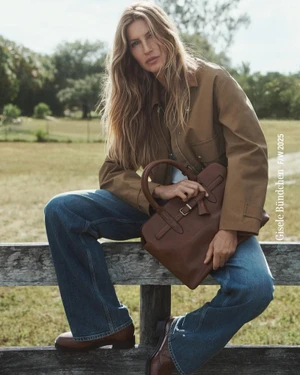 Gisele Bündchen sitting on a wooden fence wearing a brown jacket and blue jeans, holding a brown leather handbag outdoors.