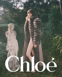 Model Grace Hartzel in a long fur coat standing next to a stone statue in a lush green garden for Chloé Winter 2025.