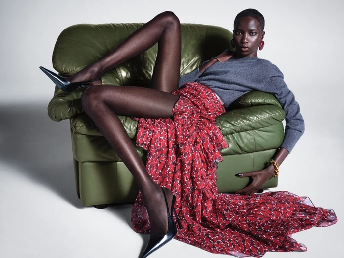 Anok Yai reclining on a green leather armchair in a red patterned skirt and grey top against a white studio background.