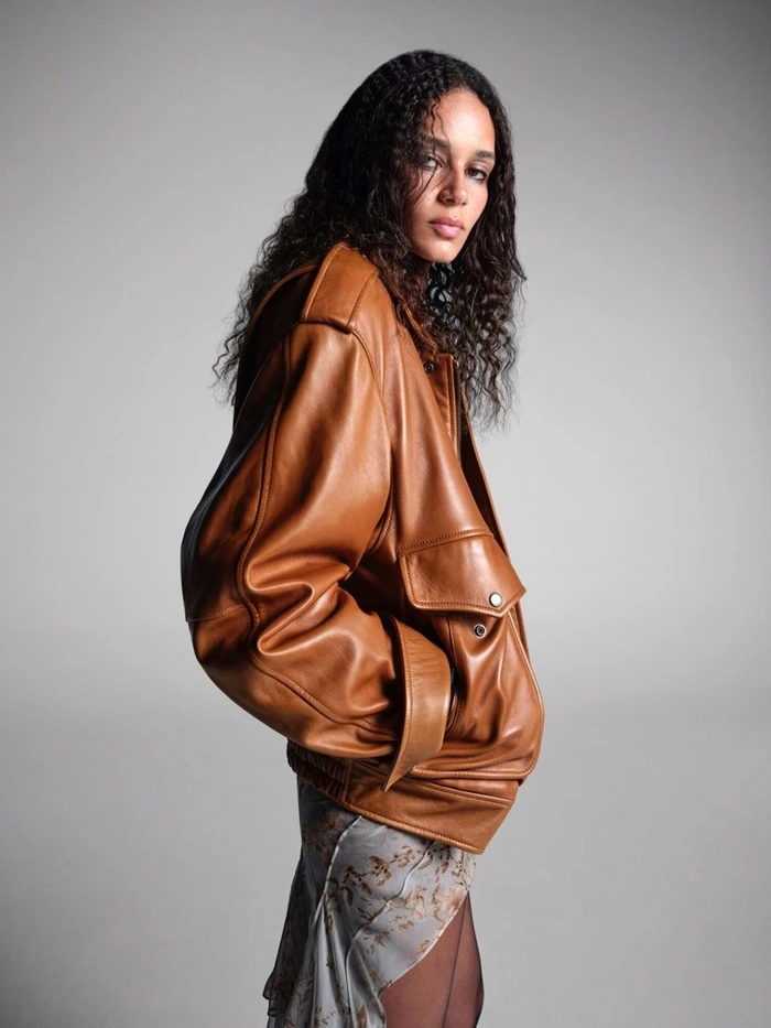 Anok Yai wearing an oversized brown leather bomber jacket against a plain grey backdrop for Yves Saint Laurent.