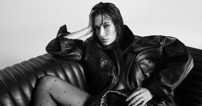 Hailey Bieber wearing an oversized black leather jacket and dotted tights seated on a leather sofa.