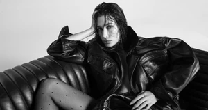 Hailey Bieber wearing an oversized black leather jacket and dotted tights seated on a leather sofa.