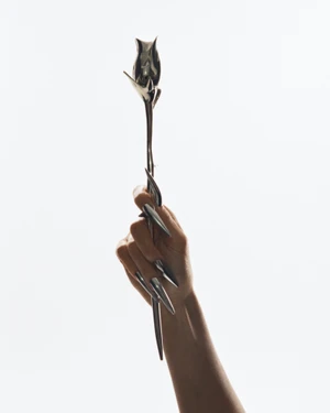 Close-up of a hand with metallic silver stiletto nails holding a sleek, sculptural metal rose against a plain white background.