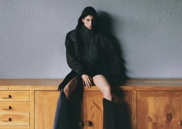 Model wearing dark jacket and tall boots sitting on a wooden cabinet against a textured grey wall
