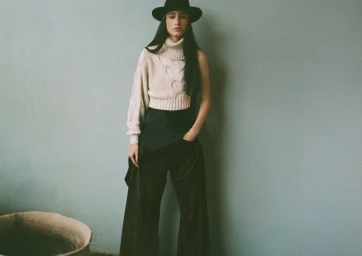 Model wearing a black wide-brim hat, cream turtleneck sweater, and dark trousers standing against a textured pale green wall.