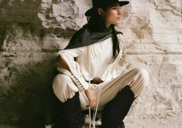 Model crouching against a textured stone wall wearing a white outfit, dark hat, and tall boots for H&M x Lorena Saravia