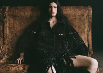 A woman with long dark hair wearing a dark fringe jacket seated against a rustic textured leather backdrop.