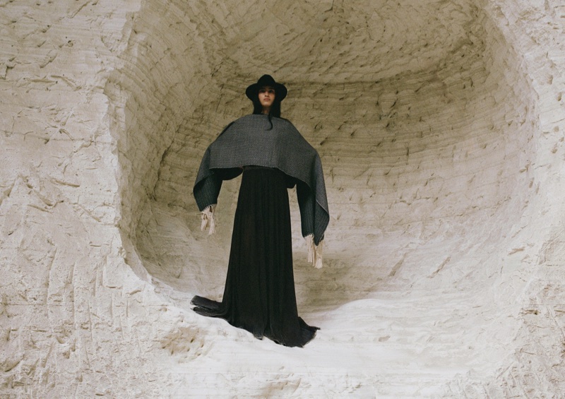 Model wearing a black hat, dark patterned poncho, and long black dress standing inside a carved white stone cave.