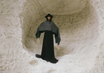 Model wearing a black hat, dark patterned poncho, and long black dress standing inside a carved white stone cave.
