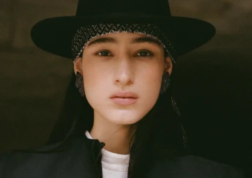 Close-up portrait of a model wearing a wide-brimmed black hat with patterned band and dark jacket.