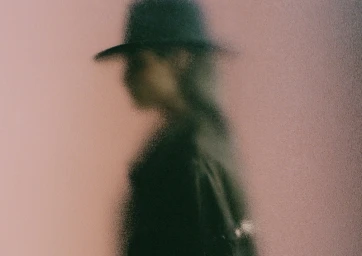 A silhouetted model wearing a fedora hat viewed through a heavily frosted or textured glass panel.