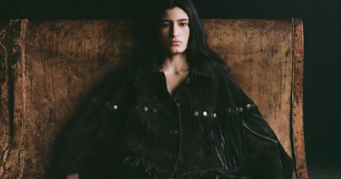 Model with long dark hair wearing a dark fringed leather jacket seated against a distressed leather background.