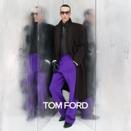 Model in sunglasses, dark coat, purple trousers, and gloves standing against a textured mirrored background with Tom Ford branding.