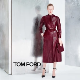 Model in burgundy textured coat holding a matching clutch standing before a reflective panel with Tom Ford branding