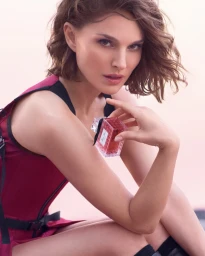 Natalie Portman holding a Miss Dior Essence fragrance bottle while wearing a dark pink and black outfit.