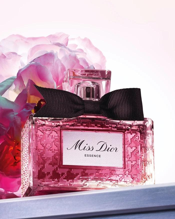 Miss Dior Essence perfume bottle with a black bow in front of pink flowers on a white background