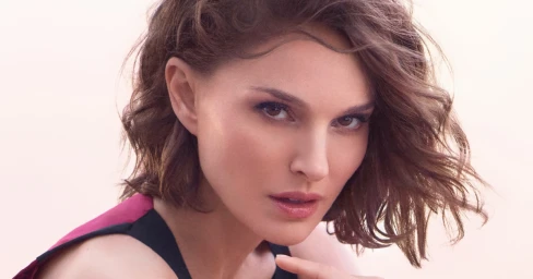 Close-up portrait of Natalie Portman with wavy brown hair, looking intently at the camera against a soft, bright background.