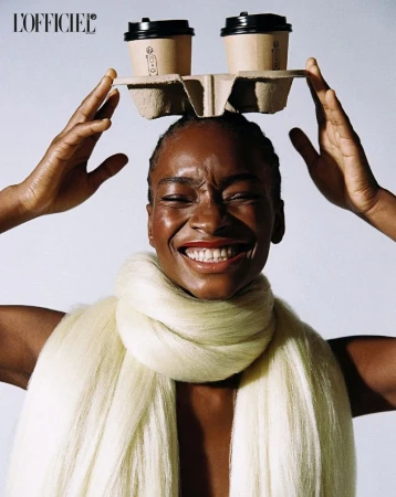 Smiling woman with short hair balances two coffee cups on her head while wearing a cream-colored scarf in a studio setting.