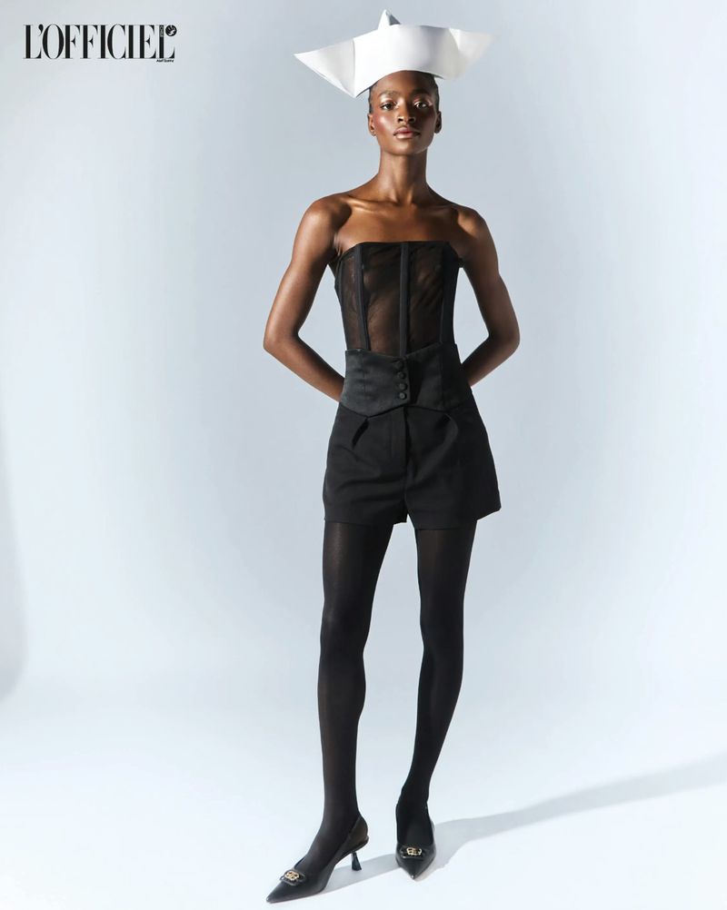 Full-length studio portrait of an adult woman wearing a corset top, shorts, tights, heels, and paper hat against a gradient white background.