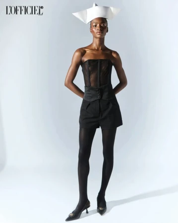 Full-length studio portrait of an adult woman wearing a corset top, shorts, tights, heels, and paper hat against a gradient white background.