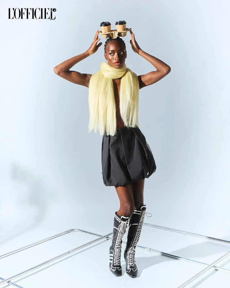 Fashion editorial shot of a Black woman balancing coffee cups on her head, wearing a yellow scarf, black skirt, and tall black boots.