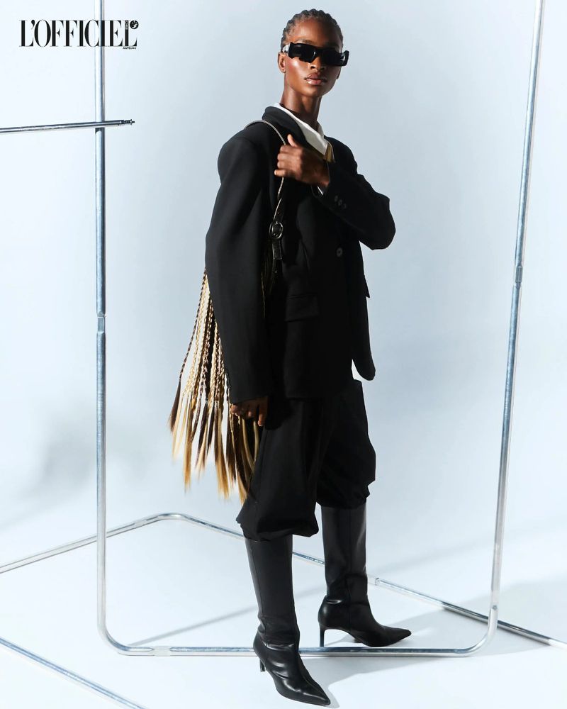 A fashion shot showcases a Black female model wearing an oversized black blazer, pants, boots, and sunglasses against a white backdrop.