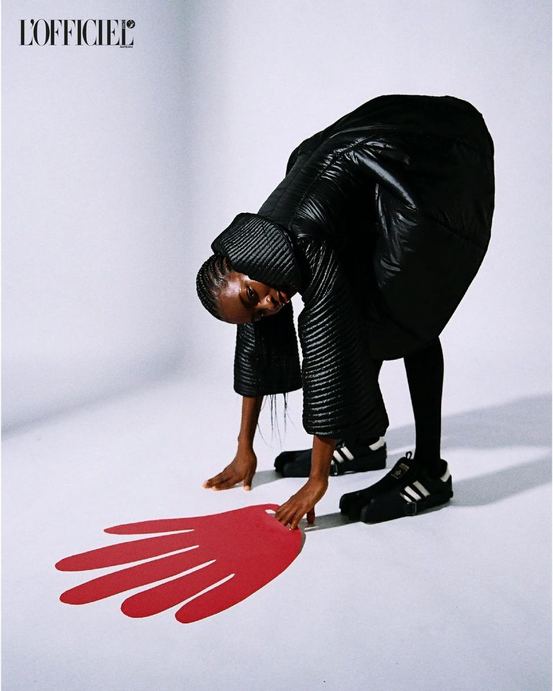 Fashion editorial shot of a Black woman posing with hands down, wearing a black puffer jacket and Adidas sneakers against a white backdrop with a red handprint.