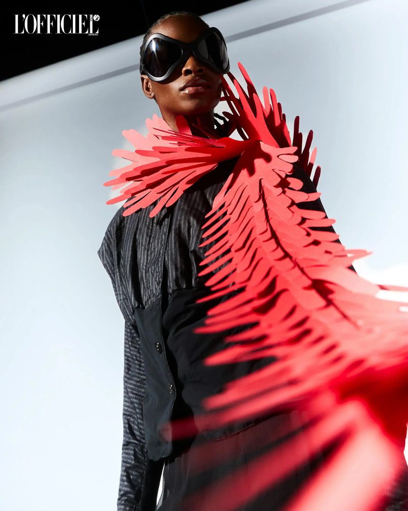 Striking fashion portrait: African female model wearing oversized sunglasses and a red paper feather collar against a neutral backdrop.