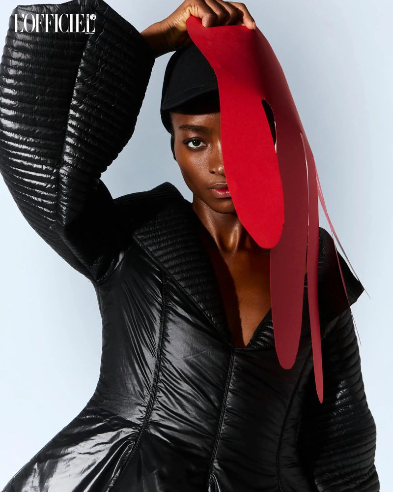 Fashionable woman poses indoors wearing a black puffer coat and cap holding a large red cutout shape that partially covers her face.